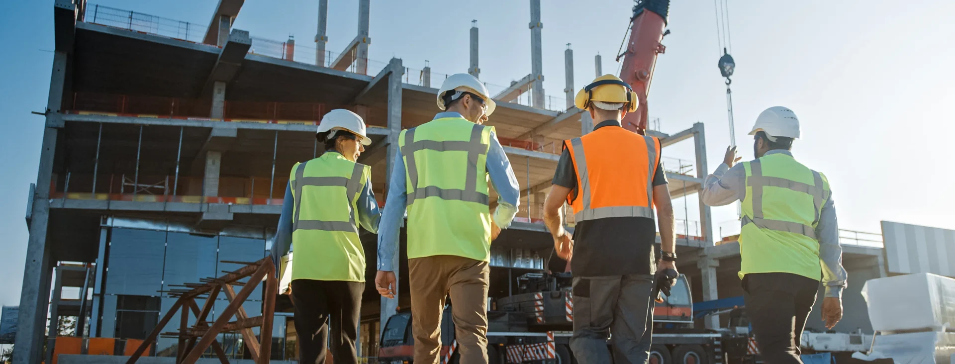 Construction workers wearing safety vests and helmets walk together at an active building site with scaffolding and cranes in the background.