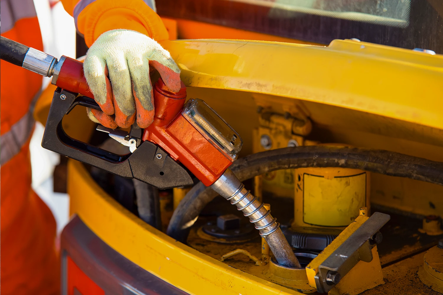 Close-up of a person wearing gloves refueling heavy machinery with a red fuel nozzle at a gas station.