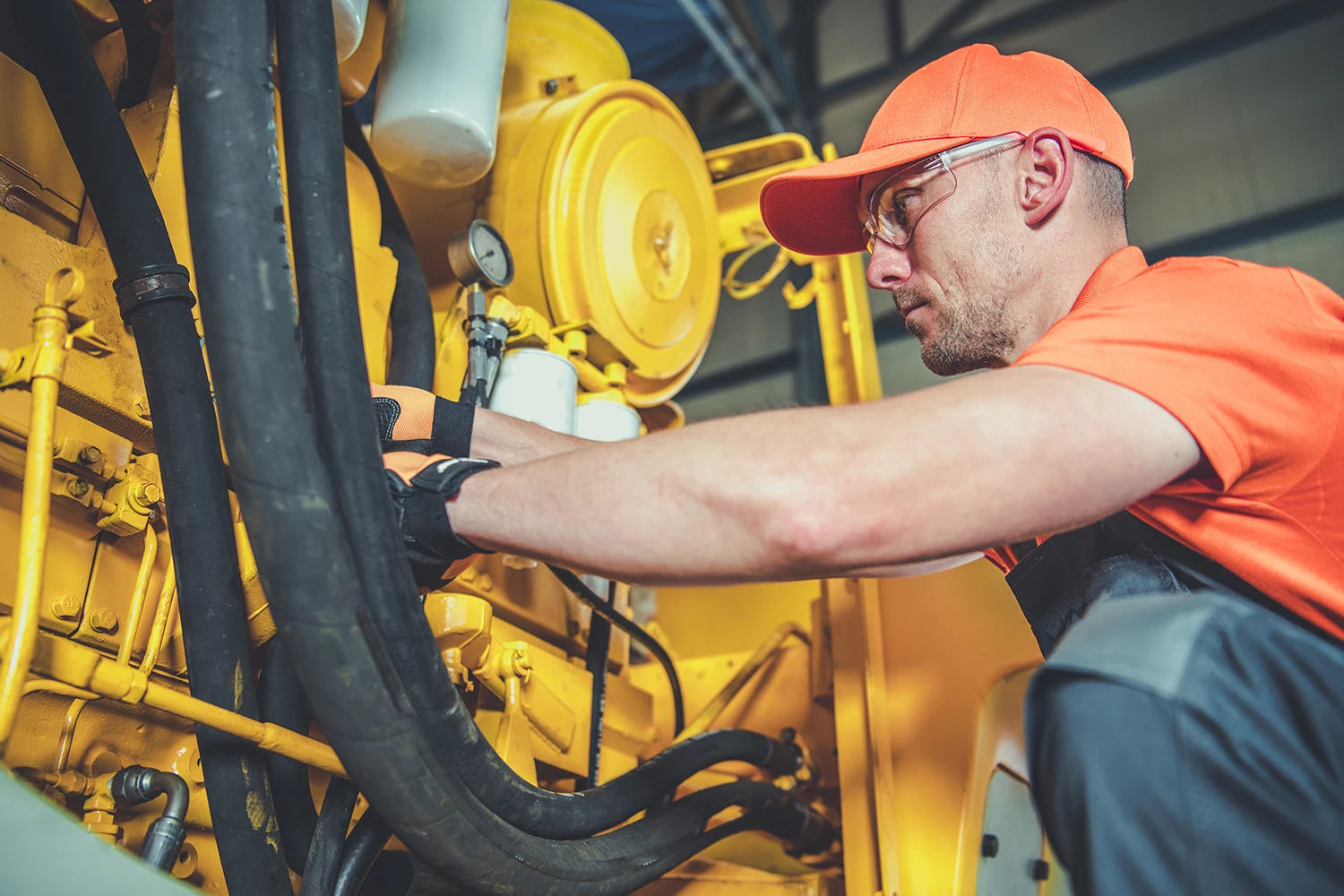 Technician wearing an orange cap and shirt inspecting industrial machinery with yellow components and black cables.
