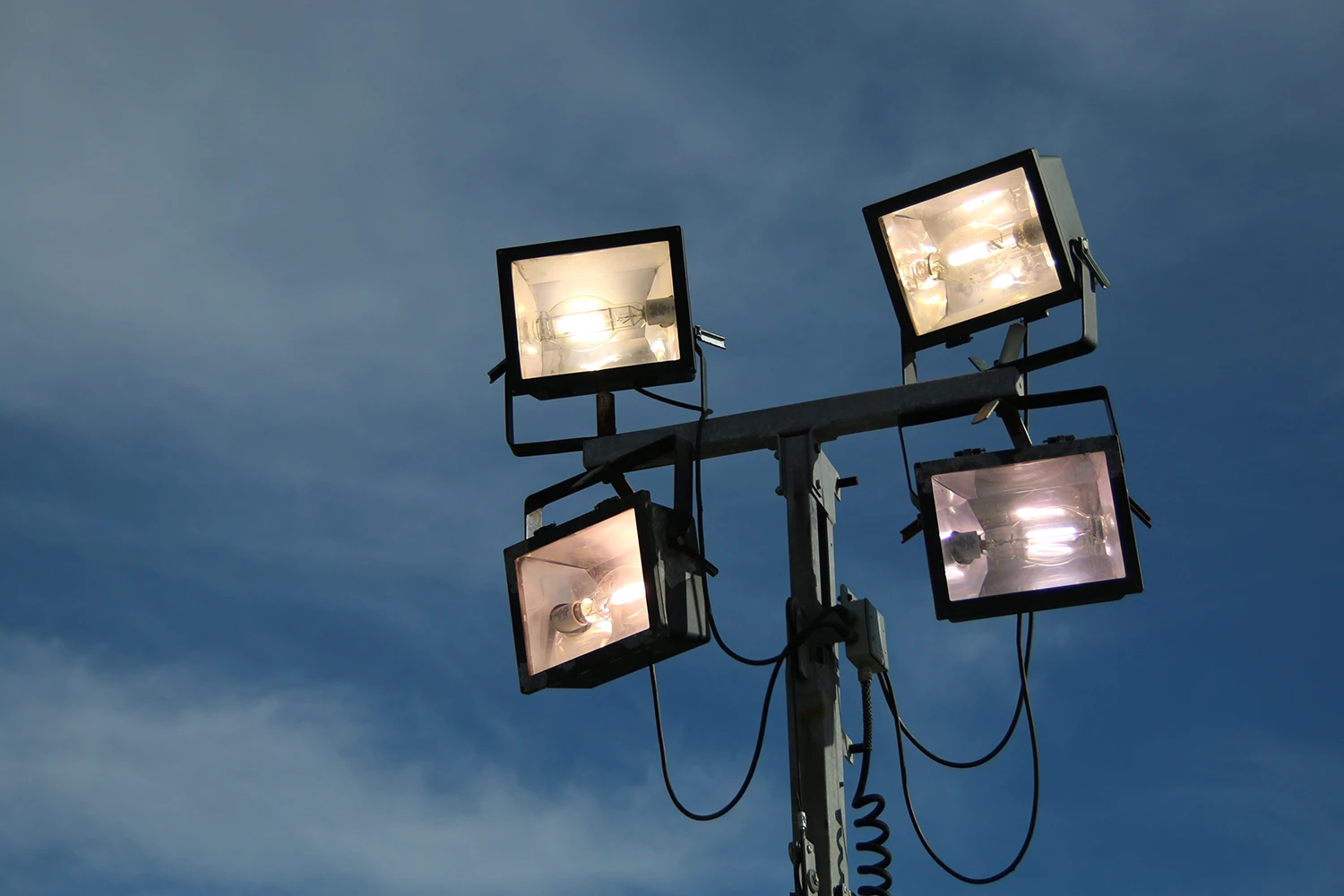 Four large outdoor floodlights mounted on a tall metal pole against a cloudy sky background.