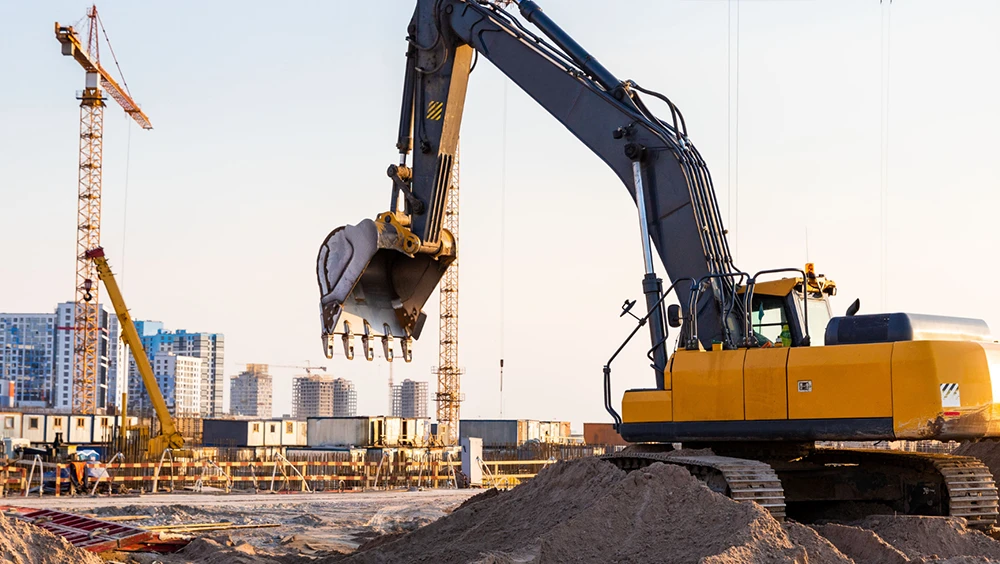 Yellow excavator lifting soil at a construction site with cranes and city buildings in the background.