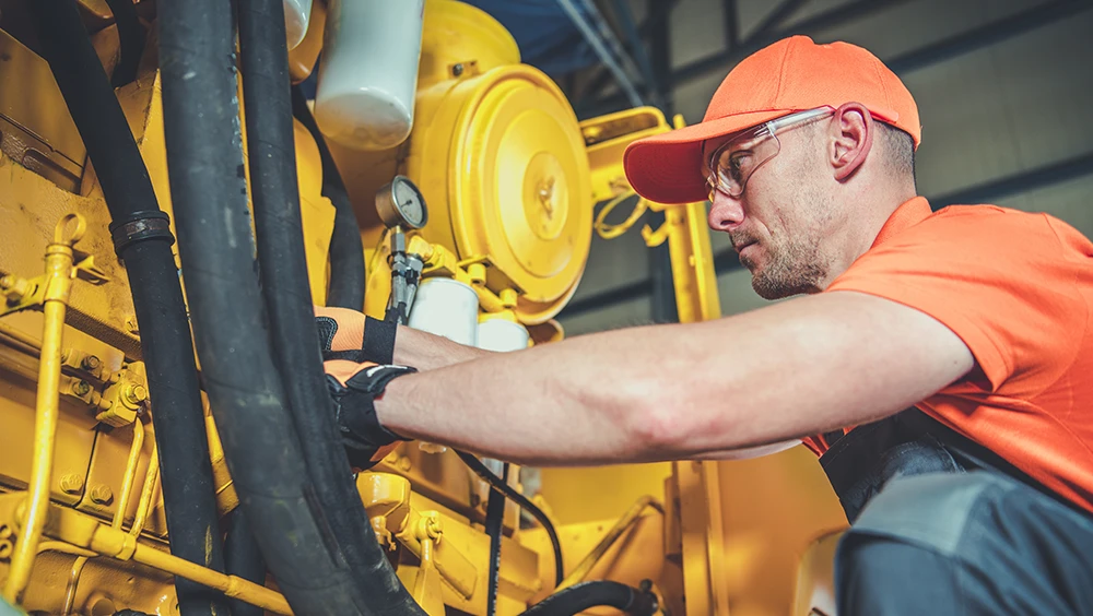 Man wearing an orange shirt and cap working on heavy machinery with yellow components in an industrial setting.