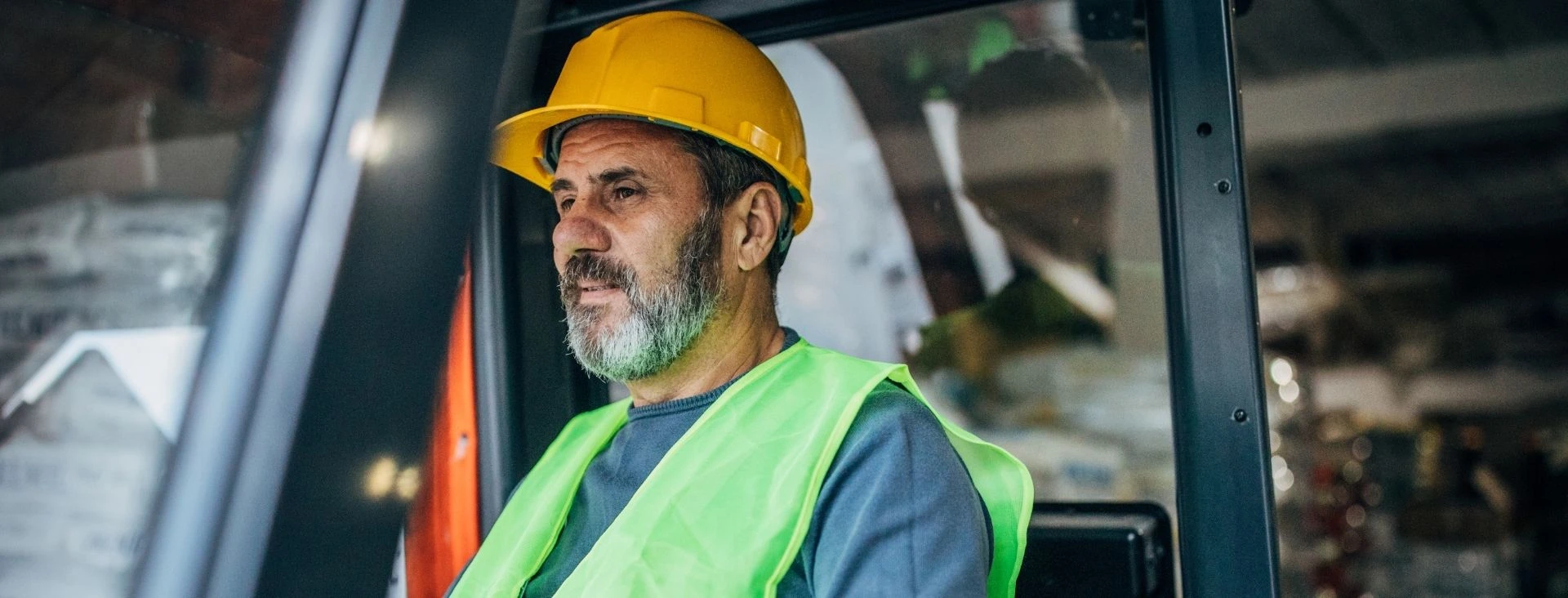 Middle-aged construction worker wearing a yellow hard hat and green safety vest operating machinery indoors.