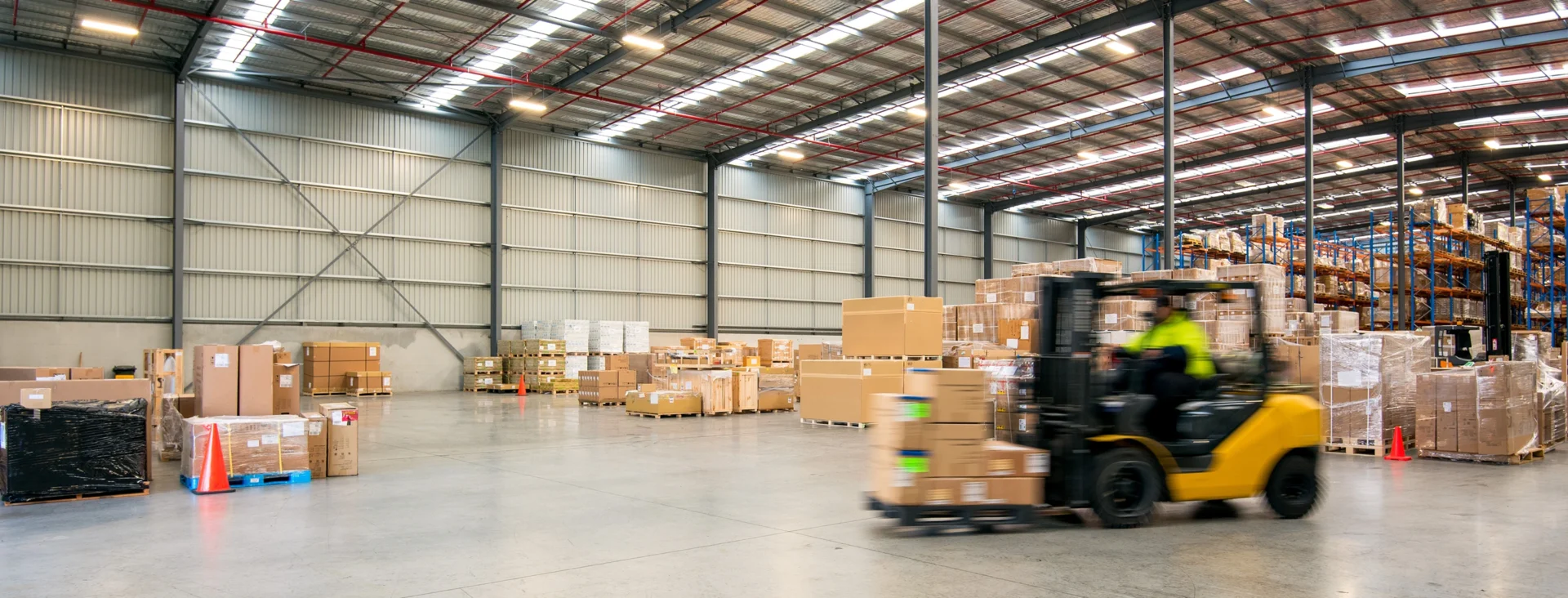 Forklift moving stacked cardboard boxes inside a large, well-organized warehouse with high shelves.