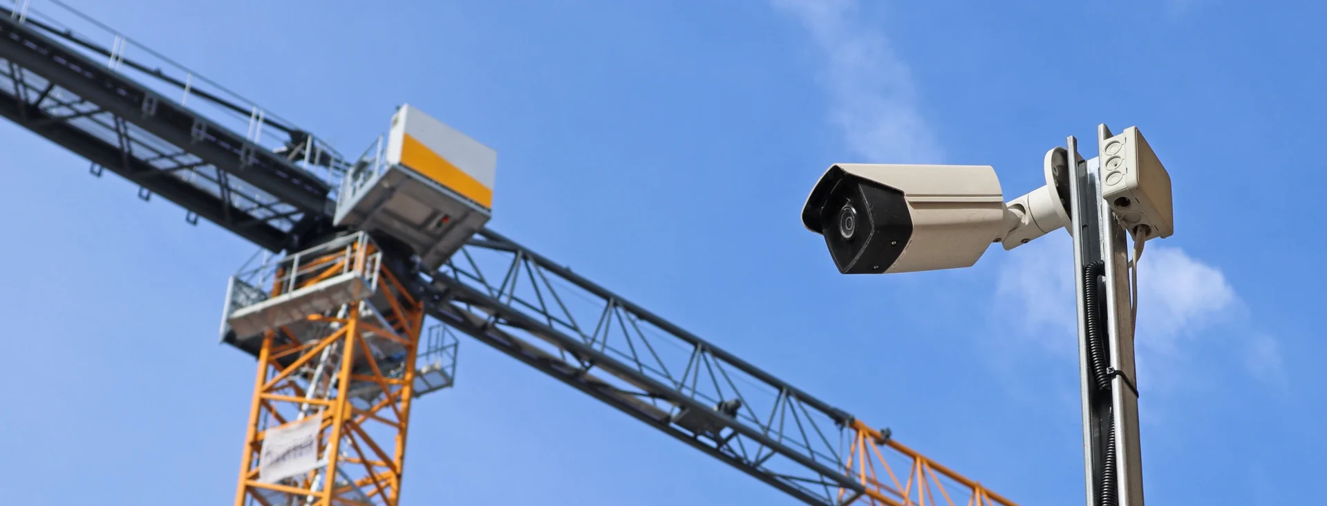 Construction crane lifting materials near a security camera mounted on a pole against a clear blue sky.