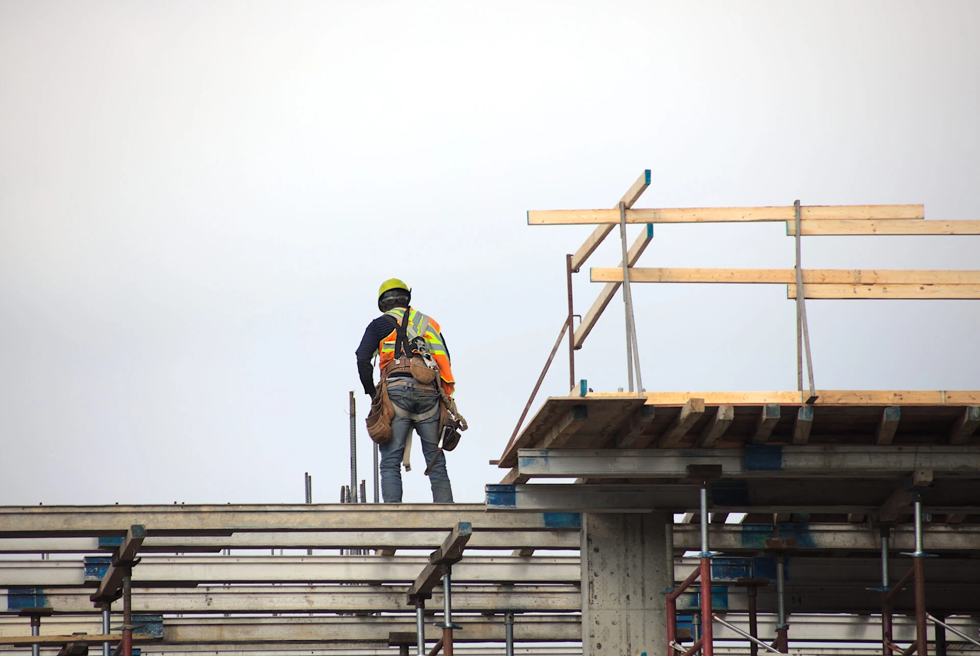 Construction worker wearing safety gear standing on scaffolding at a building site under a cloudy sky.