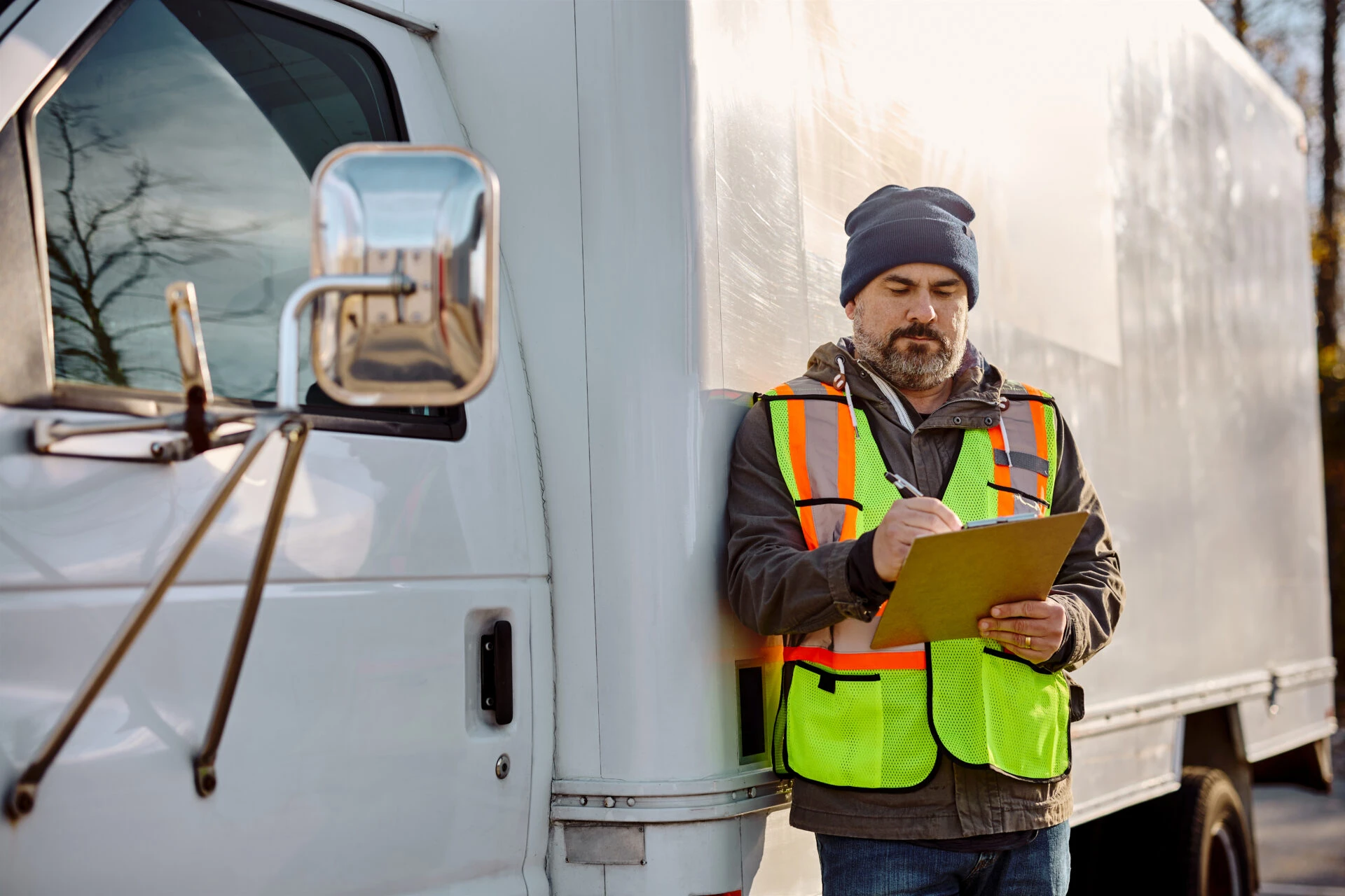 Man wearing a safety vest and beanie standing next to a white delivery truck while writing on a clipboard.