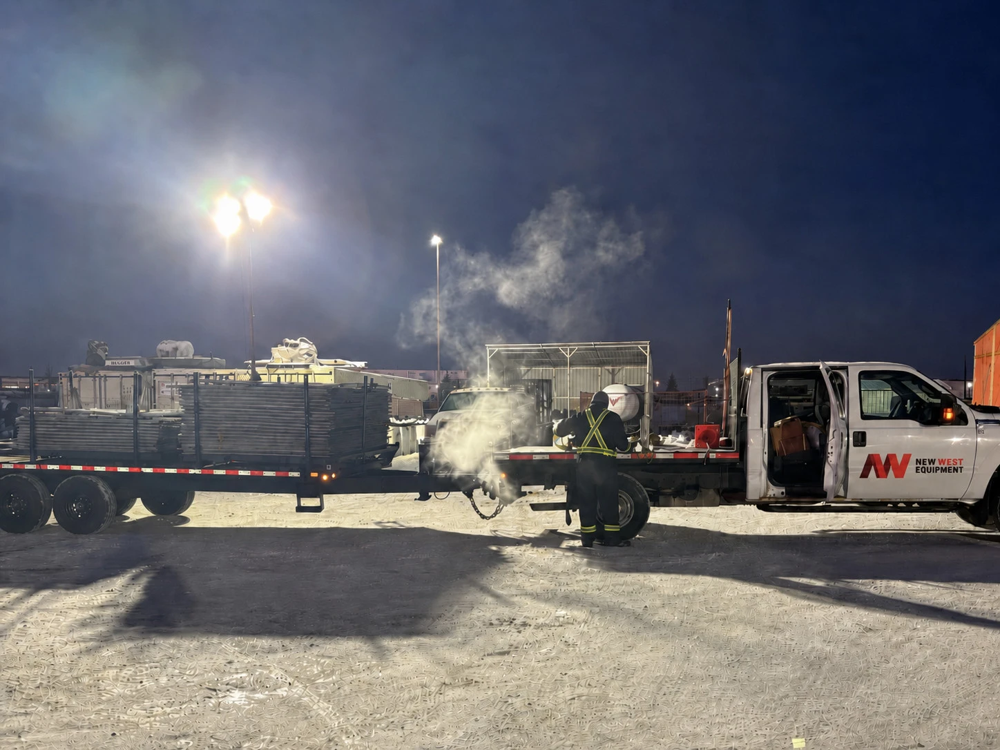 Nighttime image of a work truck with a trailer carrying equipment, emitting steam or smoke on a snowy surface.
