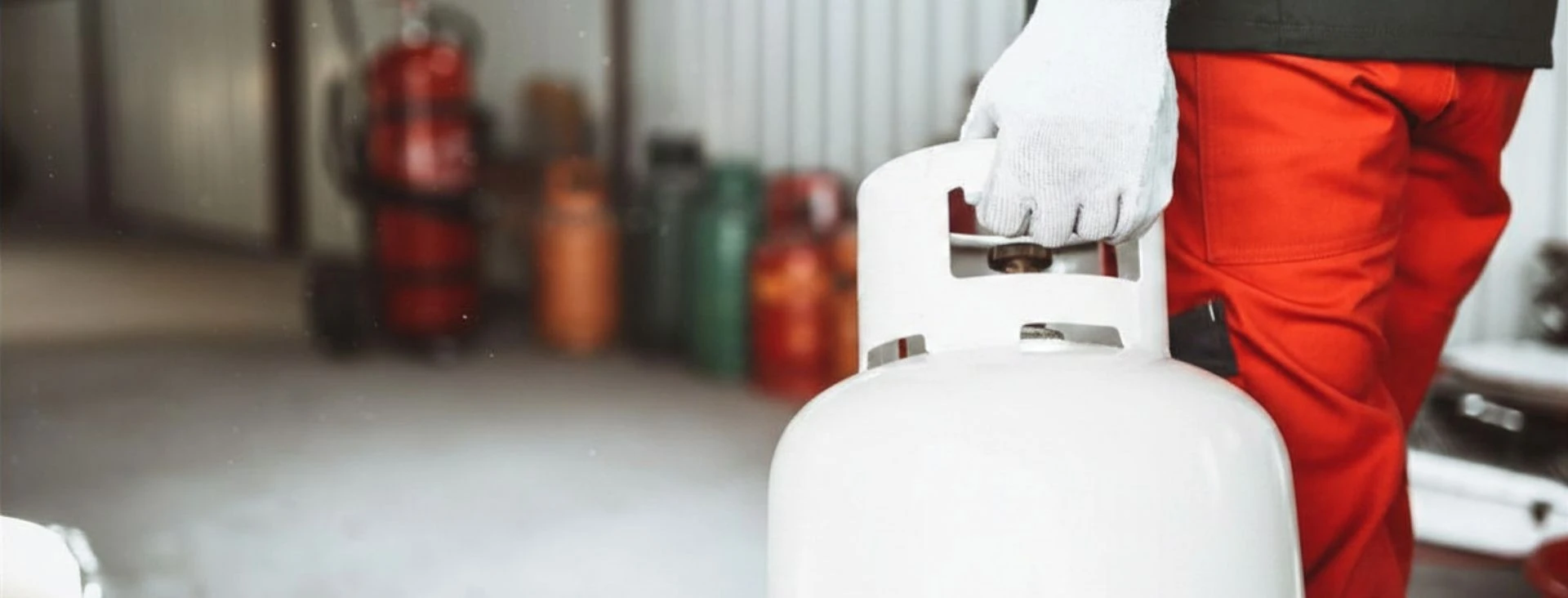 Technician in red uniform holding a white propane gas cylinder in an industrial setting.
