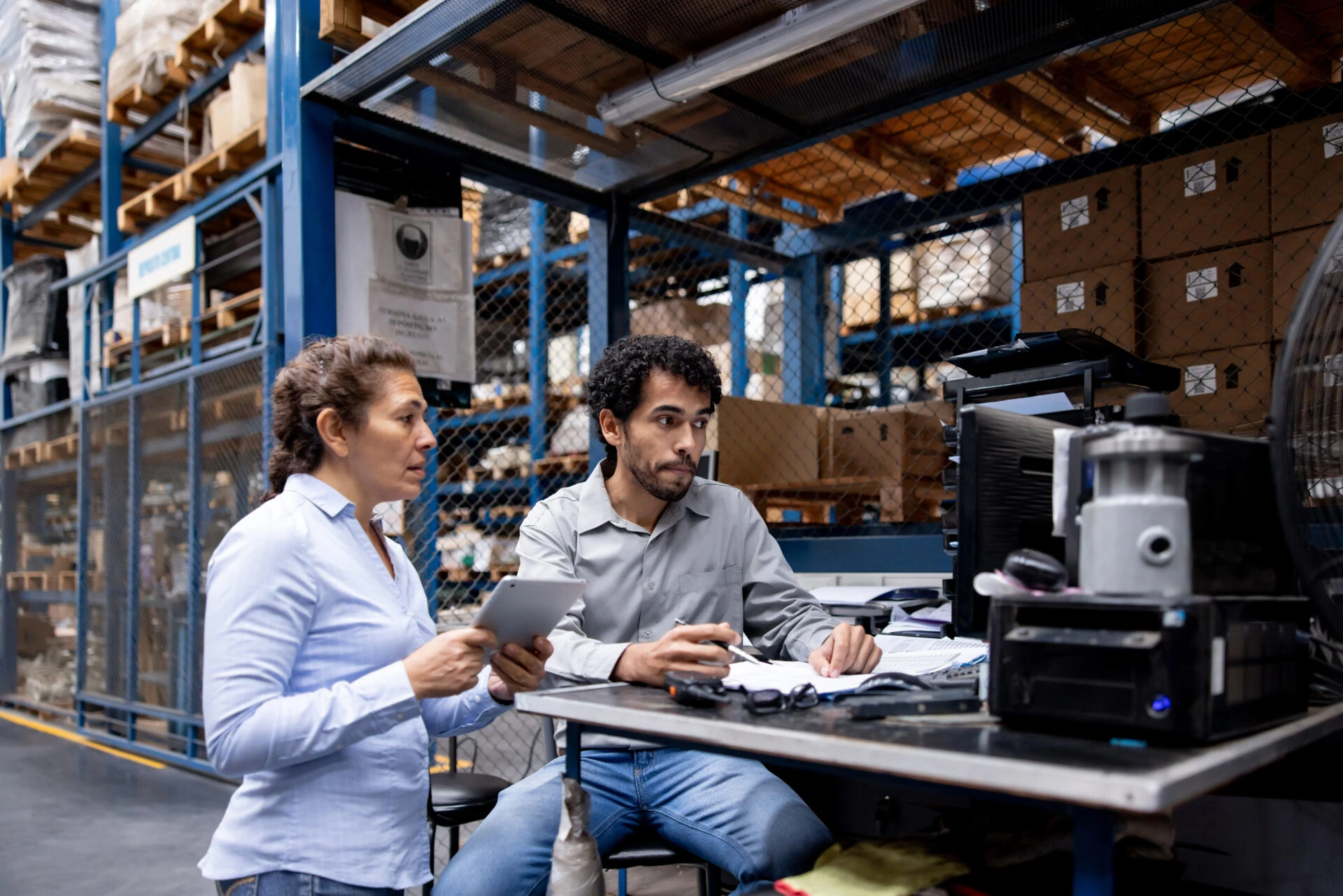 Two warehouse workers reviewing documents and using a computer at a desk in an industrial storage facility.