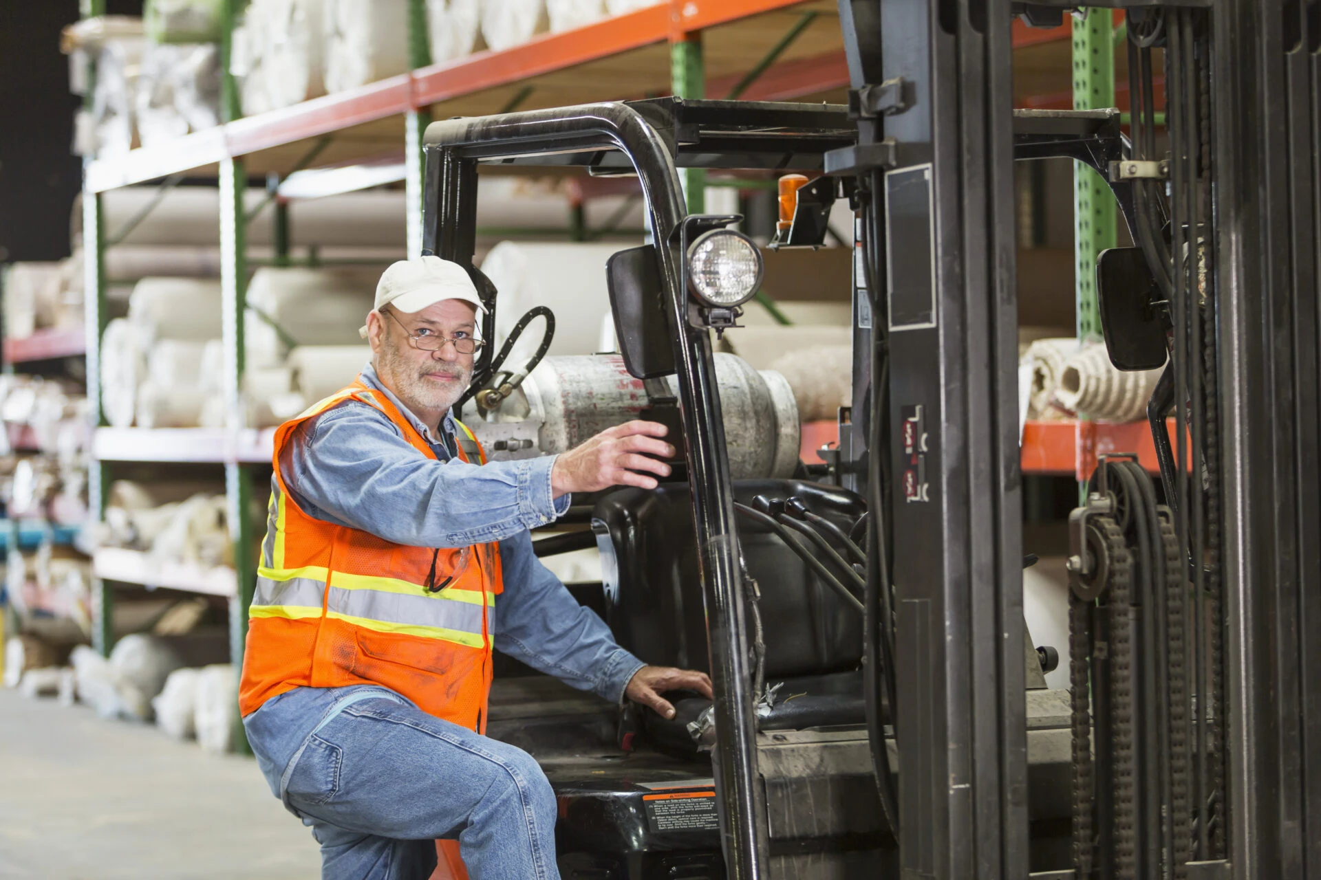 Warehouse worker in safety vest operating a forklift surrounded by stacked inventory shelves.