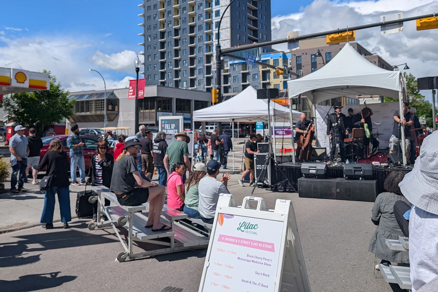 Outdoor community event with a live band performing on stage under white tents, surrounded by seated and standing attendees in a city setting.