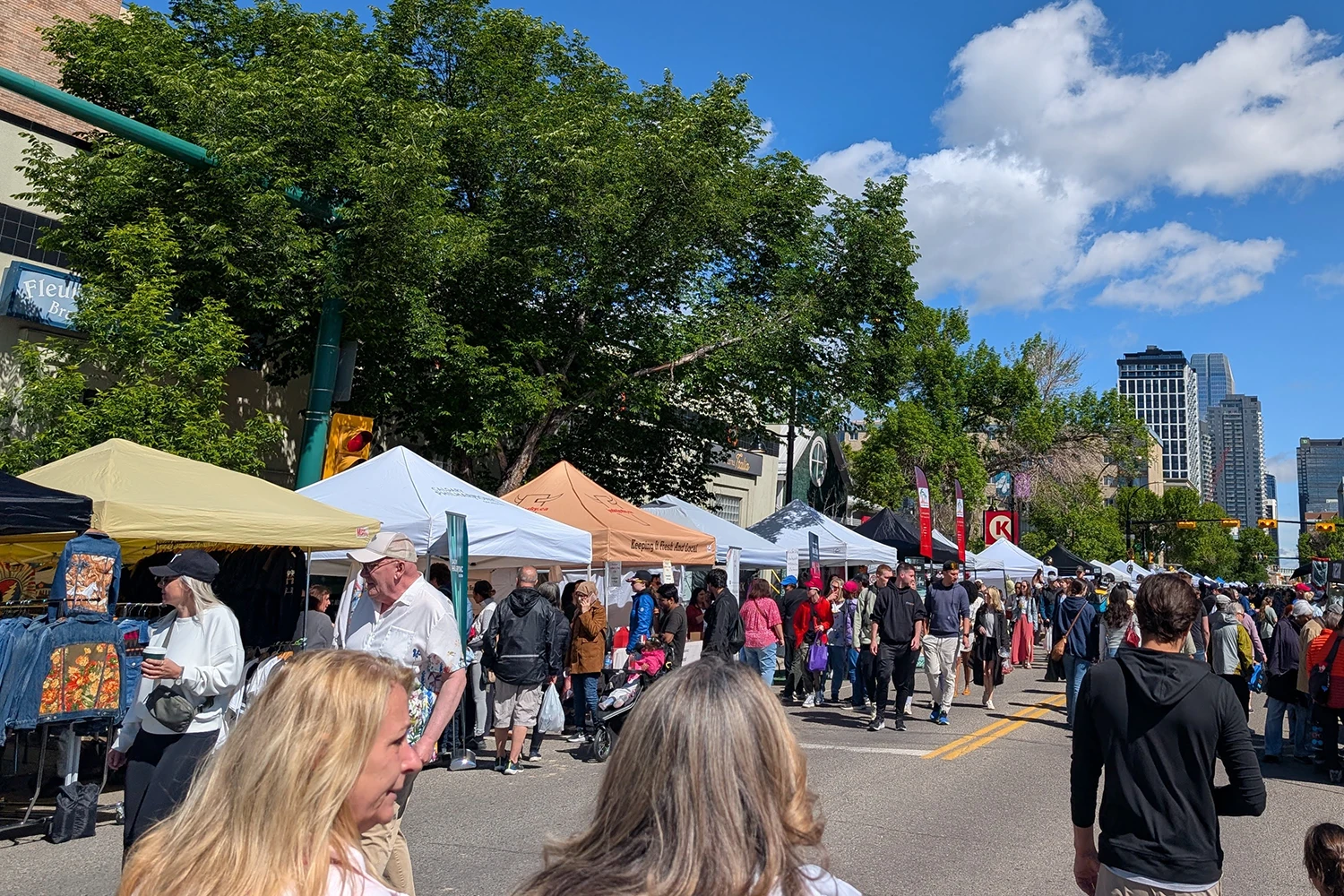 Busy outdoor street market with white and orange vendor tents and a crowd of people browsing under a partly cloudy sky.