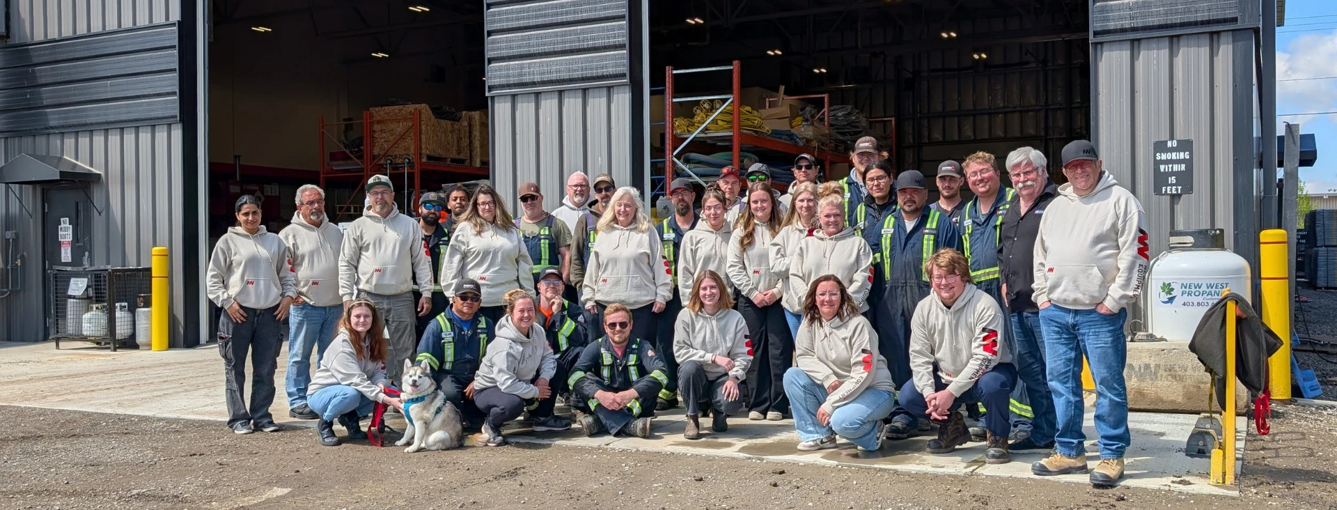 Group photo of a diverse team of people posing outside an industrial building on a sunny day.