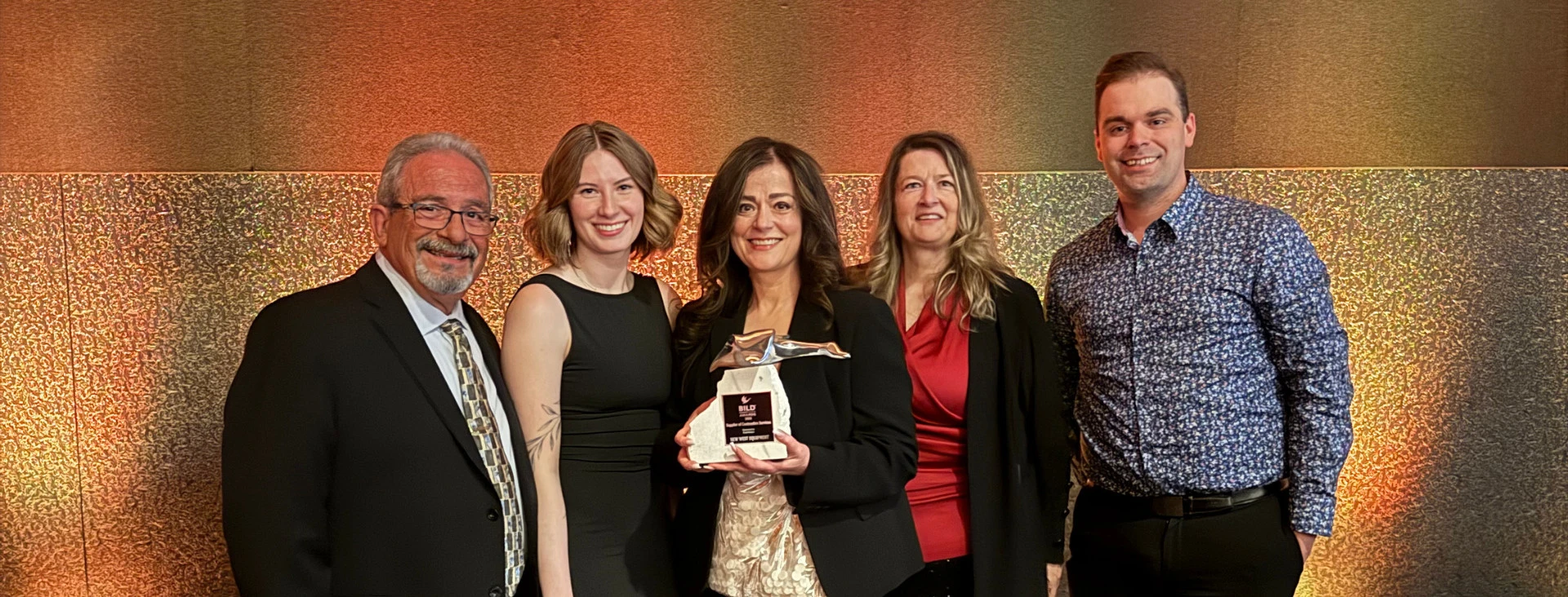 A group of five professionally dressed people standing together smiling in front of a textured brown and gold background, with the woman in the center holding an award.