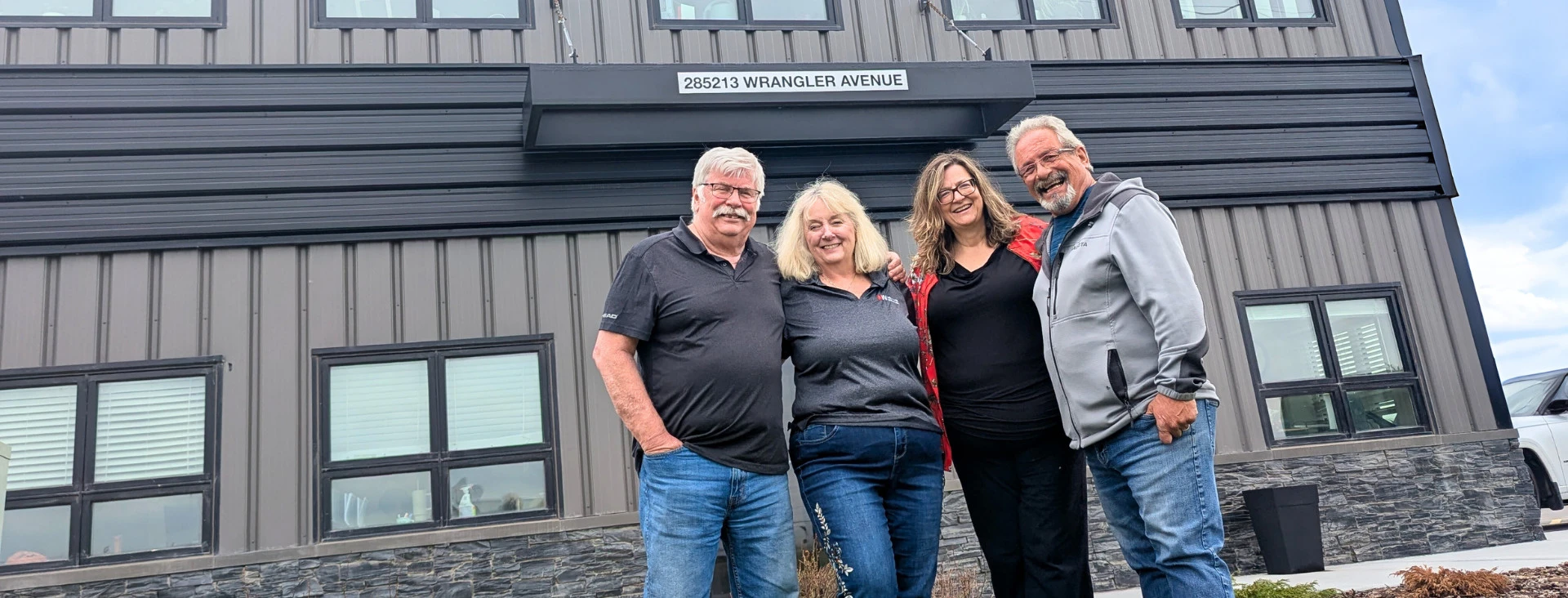 Four adults smiling and posing in front of a modern building with large black-framed windows and dark siding.