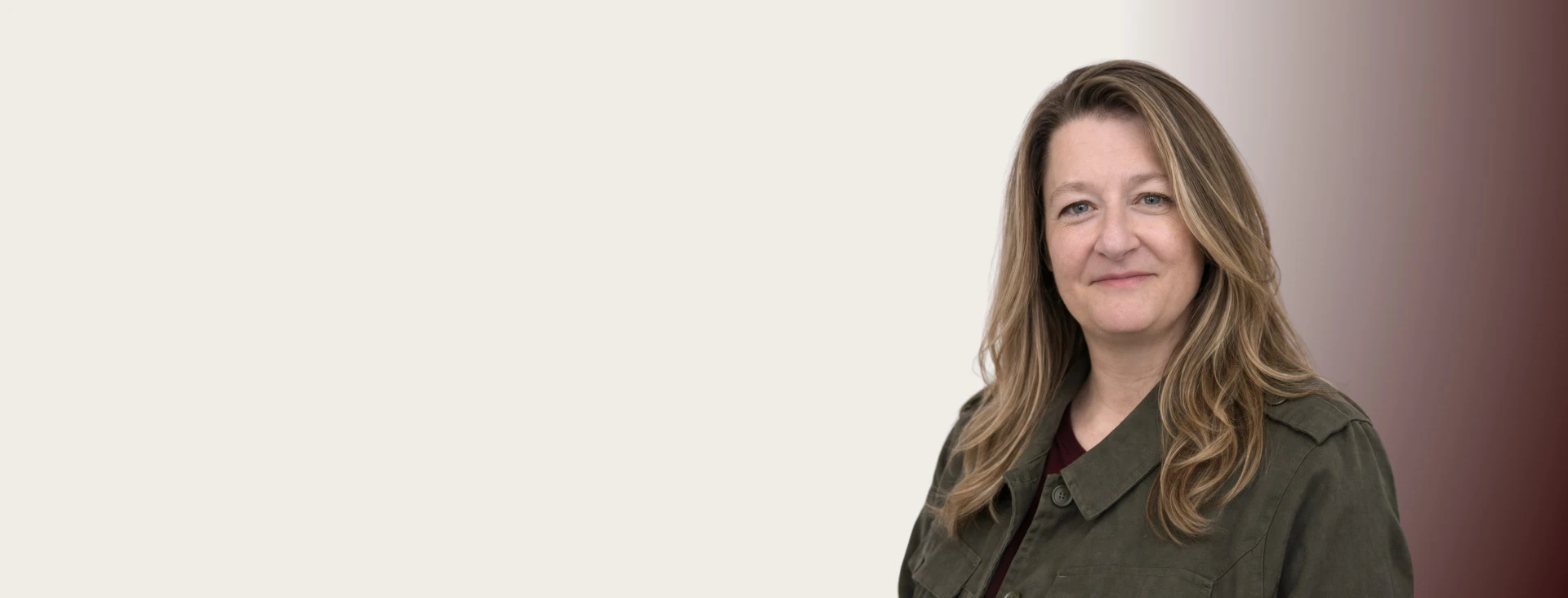 Portrait of a smiling woman with long, wavy hair wearing a dark green blazer against a plain beige background.